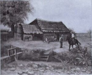 Black and white image depicting San Antonio's first settlers. There is an acequia in the foreground to the left with a lever for grabbing water out of the acequia. An old wood cabin is in the background with three settlers and two donkeys.