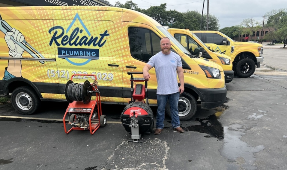 Reliant Plumbing CEO Max hicks standing in front of 3 reliant vans in yellow. His arm is resting on a hydro jetter and next to that is a plumbing camera designed to go into pipes.
